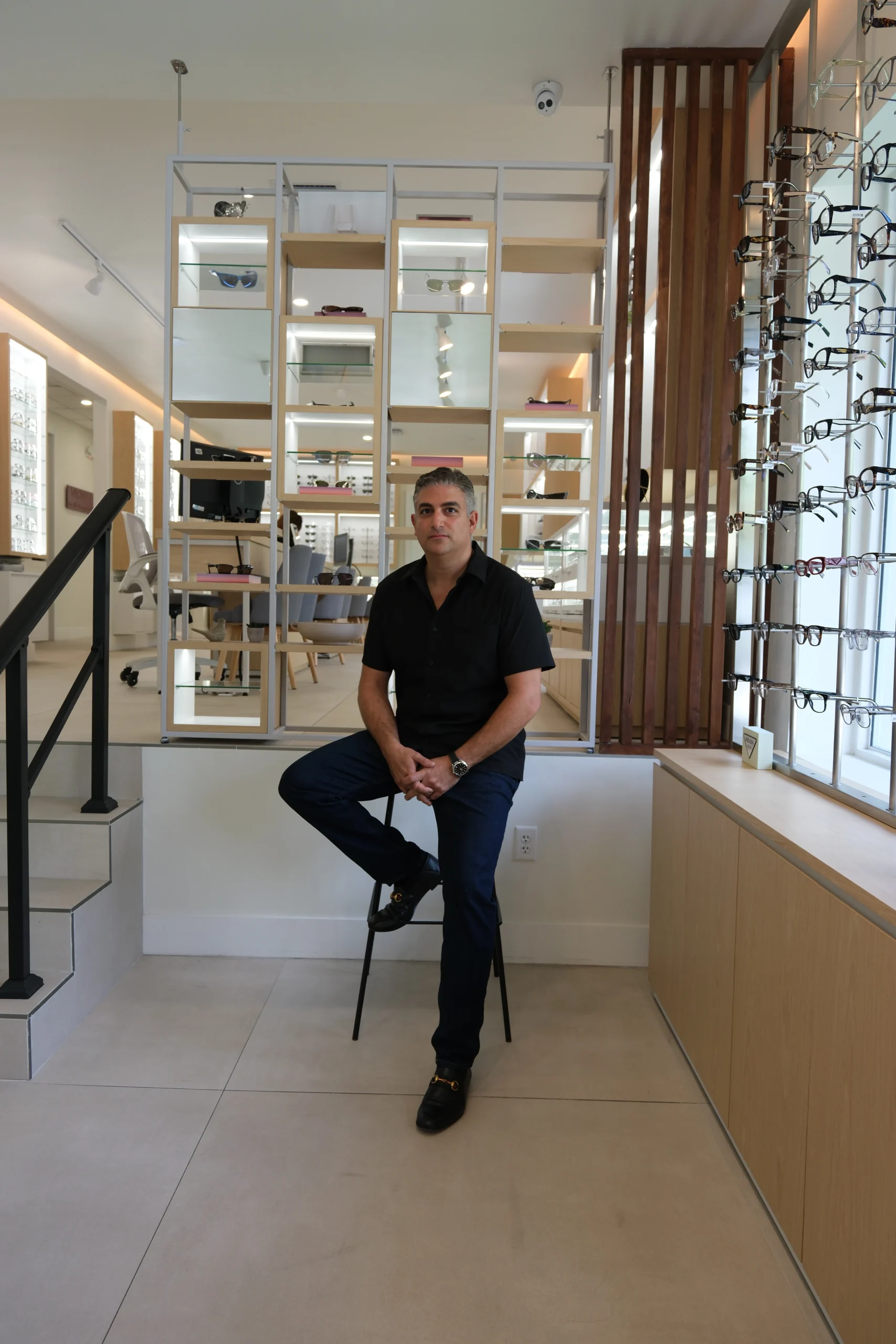 Man in a black shirt sits on a stool inside an eyewear store, with glass display shelves behind him showcasing sunglasses and frames.