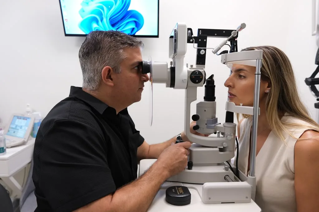 Optometrist uses a slit-lamp to examine a patient’s eyes in a clinical setting with a monitor in the background.